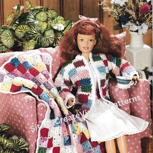 Könnte beinhalten: Eine Puppe, die eine mehrfarbige gehäkelte Jacke und ein weißes Kleid trägt, sitzt auf einem rosa Stuhl mit einer mehrfarbigen gehäkelten Decke. Die Puppe hat rote Haare und hält eine kleine weiße Tasse.