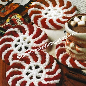 May include: Two red and white crocheted peppermint candy coasters. The coasters are on a wooden surface with a cinnamon stick and a mug of hot chocolate with marshmallows.