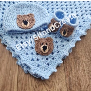 Puede incluir: Una manta de bebé de ganchillo azul claro con un gorro y zapatitos a juego. El gorro y los zapatitos tienen un aplique de oso marrón.