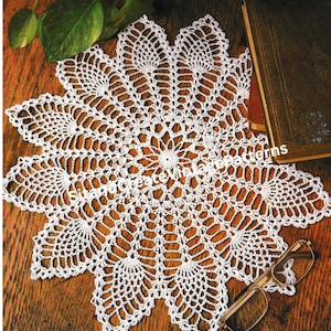 Puede incluir: Un tapete blanco hecho a mano con ganchillo con un diseño floral. El tapete es circular con un patrón calado detallado. El tapete está sobre una superficie de madera, con un libro y gafas en el fondo.