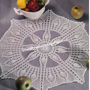 Puede incluir: Un mantel de encaje blanco con un delicado diseño floral. El mantel está colocado sobre una superficie gris con un tazón de fruta y manzanas a cada lado.