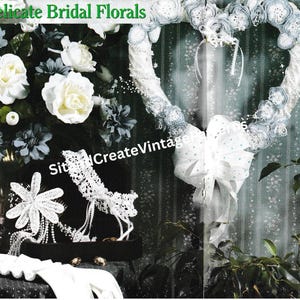 Puede incluir: Una exhibición de arreglos florales y accesorios nupciales. La imagen presenta una corona blanca en forma de corazón con un lazo grande, flores blancas y una caja negra con una flor y una liga. El texto "Delicate Bridal Florals" está en la parte superior.