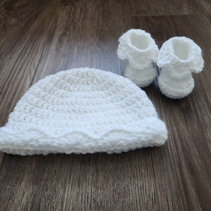 Puede incluir: Un conjunto de gorro y patucos de bebé de ganchillo blanco. El gorro tiene un borde festoneado y los patucos tienen un borde con volantes.