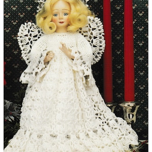 Puede incluir: Una muñeca de ángel de crochet blanco con cabello rubio y un halo de encaje blanco. La muñeca lleva un vestido de crochet blanco con un borde de encaje. La muñeca está de pie sobre un fondo estampado verde y rojo.