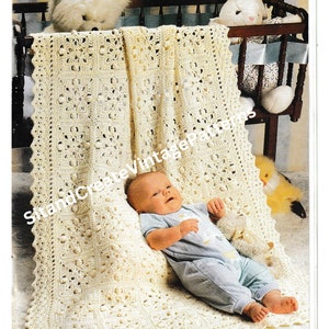 Puede incluir: Una manta de bebé de ganchillo de color crema con un patrón floral, colocada sobre una cuna. Un bebé con un mono azul está acostado sobre la manta. La imagen incluye animales de peluche y el texto "Sit and Create Vintage Patterns."