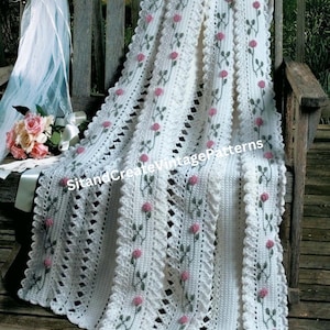 May include: White crochet baby blanket with a delicate lace border and pink rose embroidery. The blanket is draped over a wooden bench.