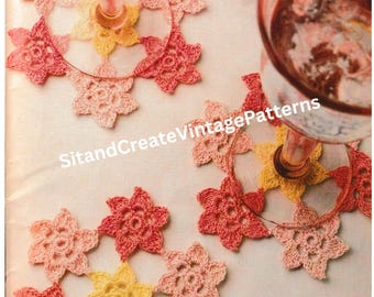 May include: Close-up of crocheted coasters in shades of pink, peach, and yellow. The coasters are made of delicate, lacy stitches and feature a flower design. The coasters are arranged on a white surface with a glass of wine in the background.