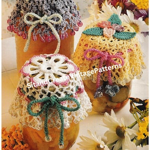 Peut inclure: Trois couvercles de bocaux en crochet avec des motifs floraux et des nœuds. Les couvercles sont dans des tons de rose, jaune et blanc. Les bocaux sont remplis de fruits conservés.