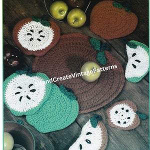 May include: Crocheted apple-shaped hot pads in green, white, and brown. The hot pads are arranged on a wooden surface with a bowl of apples in the background. The text "HandCreateVintagePatterns" is visible in the image.