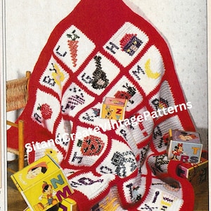 May include: A handmade, red and white crocheted blanket featuring various embroidered images within diamond-shaped blocks. The blanket displays a carrot, a violin, a strawberry, and other objects. Toy blocks with Disney characters are placed on the blanket.