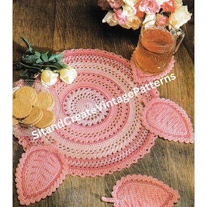 May include: A pink crocheted doily with a matching set of three pink crocheted coasters. The doily and coasters are on a wooden table with a bouquet of pink and white roses in a glass vase.