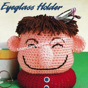 Puede incluir: Portagafas de ganchillo con forma de personaje de dibujos animados sonriente con pelo marrón, piel rosa y camisa roja. El portagafas tiene una ranura para las gafas en la parte superior de la cabeza.