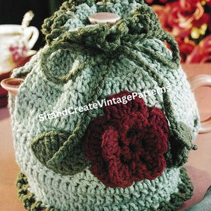 Può includere: Un copriteiera in crochet di colore verde e bordeaux con un motivo a rosa. Il copriteiera ha la forma di una teiera e ha un fiocco verde in cima. Il testo "Rose Tea Cozy" è visibile nella parte superiore dell'immagine.