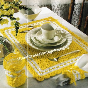 May include: A yellow and white crocheted placemat with a white napkin ring and a glass with a crocheted cover. The placemat is on a white tablecloth with a white cup and saucer on top of it.