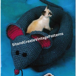 May include: A crocheted cat bed shaped like a mouse. The bed is blue with pink ears and a pink nose. A white cat is sitting inside the bed. The text "Snuggy Mouse Cat Bed" is at the top of the image. The text "SitandCreateVintagePatterns" is in the middle of the image.
