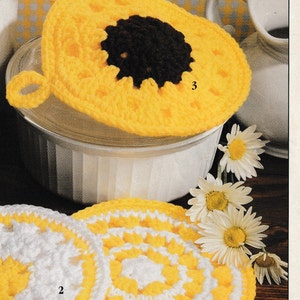 Puede incluir: Tres posavasos de ganchillo, dos son blancos y amarillos con un patrón circular, y uno es amarillo con un centro marrón que se asemeja a un girasol. Los posavasos tienen lazos para colgar.