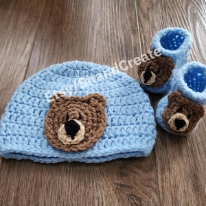 May include: A blue crocheted baby hat and booties set with brown bear appliques. The hat has a bear face on the front and the booties have a bear face on the top.