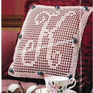 May include: A white crocheted pillow with a large "M" monogram in the center. The pillow is decorated with blue fabric rosettes and is sitting on a red velvet surface.