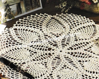 May include: A cream-coloured, circular doily with an open, lacy crochet pattern. The doily is on a dark wooden surface, with the text "Pineapples & Petals" at the top. A pen and some postcards are also visible.