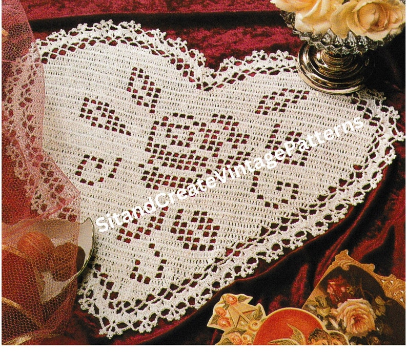 May include: A heart-shaped, off-white crocheted doily with a scalloped edge. The doily has a central design with the text "SitandCreateVintagePatterns". The doily is displayed on a burgundy surface with decorative elements.