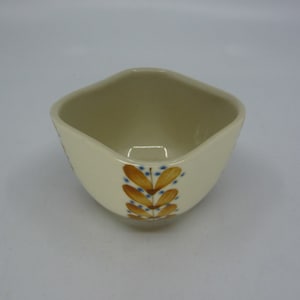 Puede incluir: Un pequeño cuenco de cerámica blanca con forma cuadrada y bordes redondeados. El cuenco tiene un diseño floral en tonos marrones y azules.