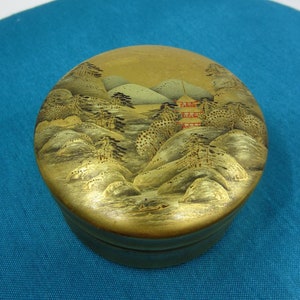 May include: A round, gold-coloured box with a Japanese landscape scene painted on the lid. The scene features mountains, trees, and a red pagoda.