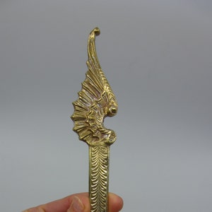 May include: A gold-colored metal letter opener with a decorative winged design at the top. The wings are detailed and intricate, and the letter opener has a long, slender blade.