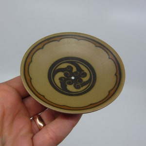 Peut inclure: Un petit plat rond en céramique brun avec un motif noir et orange. Le motif présente un motif en spirale stylisé.