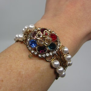 Peut inclure: Un bracelet en or avec un grand charme en forme de fleur coloré et des accents de perles blanches.