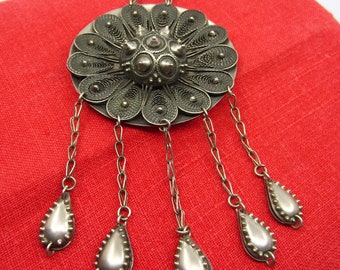 May include: A silver filigree pendant with a circular design and five teardrop-shaped charms hanging from it. The pendant is on a red fabric background.