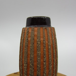 May include: A brown and orange striped ceramic vase with a white base. The vase has a textured surface and a dark brown rim.
