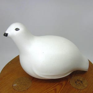 May include: A white ceramic bird ornament with black detailing on the beak and eyes. The bird sits on a round wooden base. The figurine has a smooth, matte finish and a rounded body.