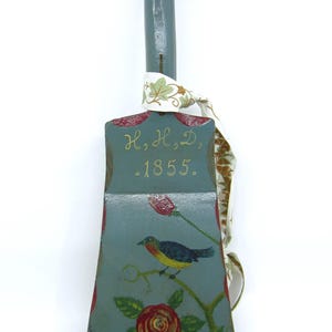 Peut inclure: Une cloche en bois vintage peinte à la main, avec une longue poignée et une base évasée. La cloche est peinte en bleu-vert avec un motif d'oiseau et de rose. Le texte "H, H, D, -1855." est peint sur la cloche. Un ruban est attaché à la poignée.