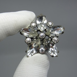 May include: A silver-toned flower brooch with clear rhinestones. The brooch is held in a white gloved hand.