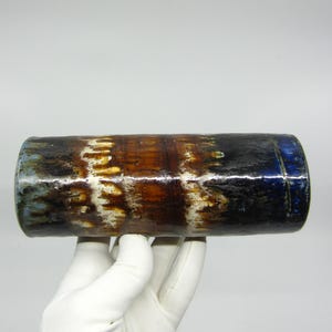 May include: A cylindrical ceramic vase with a textured surface. The vase features a blend of colors, including brown, black, white, and blue. The handmade pottery has a glossy finish and is held by a gloved hand.