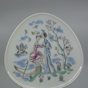 May include: A white ceramic plate with a blue and green design featuring a couple in a boat, a tree, and birds. The plate is decorated with a whimsical illustration and the text "Rygiel".