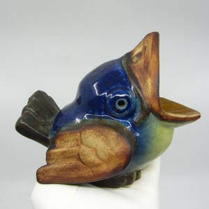 May include: A ceramic bird figurine with a wide open beak. The bird has a blue head, brown beak, and tan wings. The figurine is resting on a white surface.