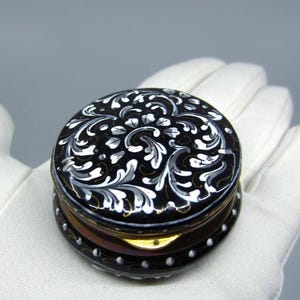 May include: A small, round, black lacquer box with a hinged lid. The lid features intricate white and grey floral scrollwork patterns. The base of the box is also decorated with a row of small white dots along the edge, and a gold-toned metal rim.
