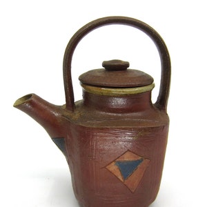 May include: A small, brown ceramic teapot with a lid and a handle. The teapot has a geometric design with blue, red, and white accents.