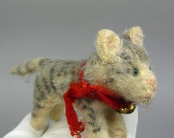 May include: A vintage stuffed cat figurine with a gray and cream-colored body and black stripes. It has a red ribbon around its neck and a small gold bell. The cat's ears are cream-colored, and it has green eyes and a pink nose.