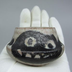 May include: A small ceramic bowl with a distinctive design. The bowl has a black and white pattern that looks like a face. The rim is reddish-brown. The bowl is held in a white glove.