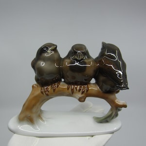 May include: A ceramic ornament featuring three brown birds on a brown branch. The birds have dark brown and grey feathers. The branch rests on a white oval base. A decorative item.