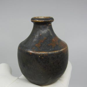 May include: A small, dark brown ceramic vase with a rounded body and a narrow neck. The vase has a speckled glaze with hints of gold.