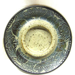 Puede incluir: Plato de cerámica con una zona central empotrada y un borde decorativo. El borde presenta un borde azul oscuro con diseños florales y animales grabados en tonos verdes y azules. El plato tiene un interior moteado de color blanquecino.