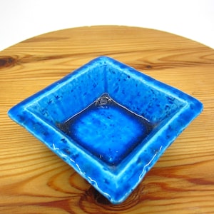 Puede incluir: Cenicero cuadrado de cerámica con un vibrante esmalte azul brillante. El cenicero tiene forma de diamante con bordes ligeramente elevados y un interior oscuro. Colocado sobre una superficie de madera.