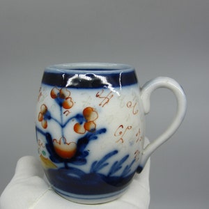 Könnte beinhalten: Eine kleine, weiße Keramiktasse mit einem blauen und roten Blumendesign. Die Tasse hat einen abgerundeten Körper und einen Henkel. Das Design beinhaltet ein Blumenmotiv mit roten Blüten und blauen Blättern.