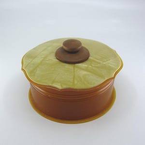May include: A vintage, round trinket box with a lid. The box is a warm brown color with a scalloped edge and a light green lid. The lid has a small, round, brown knob. The box sits on a matching base.