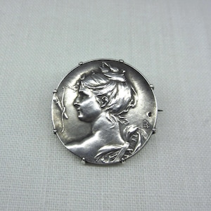 May include: A silver-toned brooch with a detailed profile of a woman's face in a circular design. The woman's hair is styled up, and the brooch has a pin on the back. The brooch is likely made of metal.