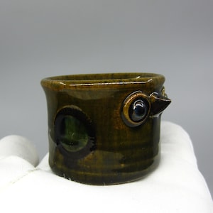 Può includere: Un piccolo porta uova in ceramica marrone con un viso fantasioso. Il porta uova ha due occhi verdi e neri e un piccolo becco.