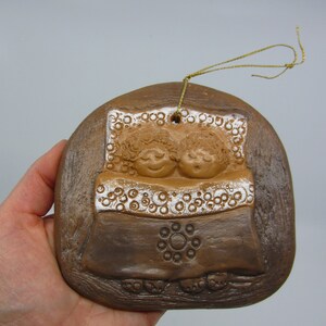 May include: A brown ceramic ornament depicting two figures sleeping in a bed with a white patterned blanket. The ornament has a gold string for hanging.